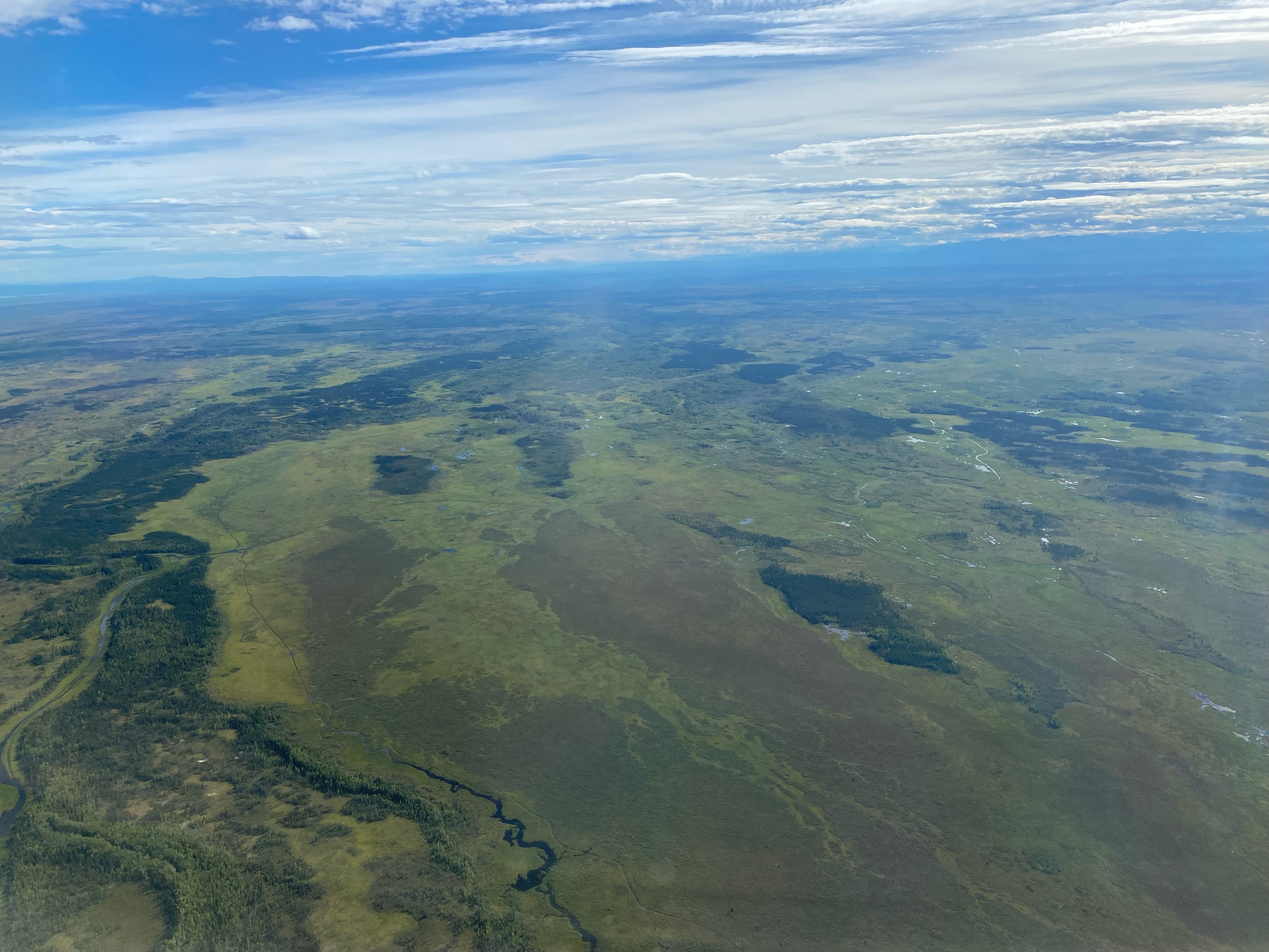 Aerial view of Alaska out the window of a NASA Gulfstream III plane. The land is mostly lush and green, with lakes dotting it and rivers snaking through the landscape. The sky is blue and has puffy white clouds.