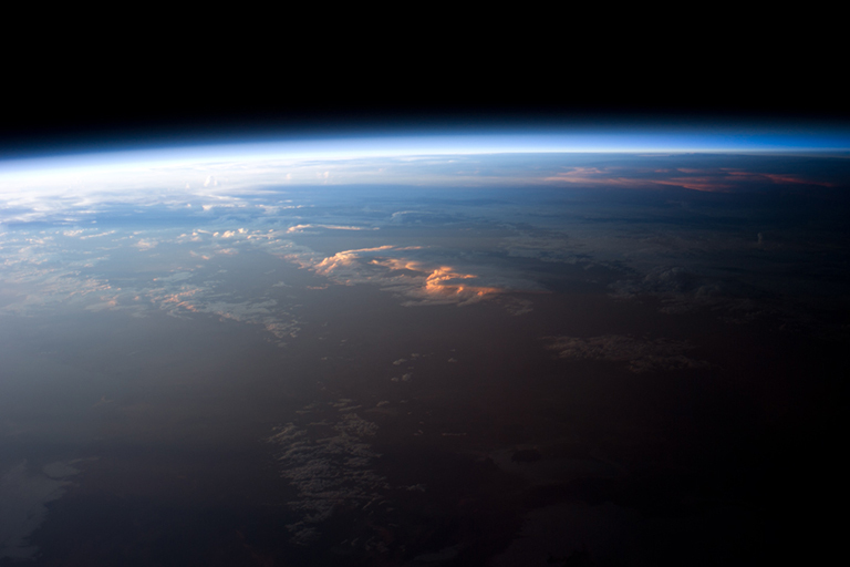 A view of central South America as seen from the International Space Station. Layers of the Earth’s atmosphere, colored bright white to deep blue, are visible on the horizon (or limb). The highest cloud tops have a reddish glow due to direct light from the setting Sun, while lower clouds are in twilight.