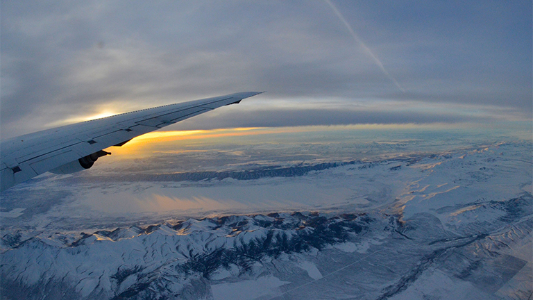A view looking out over the plane's wing and a dim sun shining on snow-covered mountains.